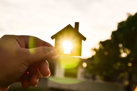 Eine Hand hält ein kleines Haus aus Holz gegen die Sonne und die Sonne scheint durch ein Fenster des kleinen Hauses.